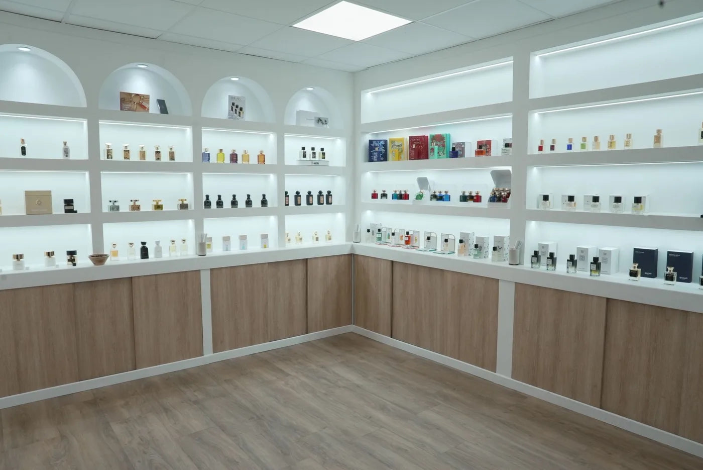 Angle du showroom AD Parfumerie : mur de niches arquées rétroéclairées, soubassement en bois clair et flacons colorés alignés
